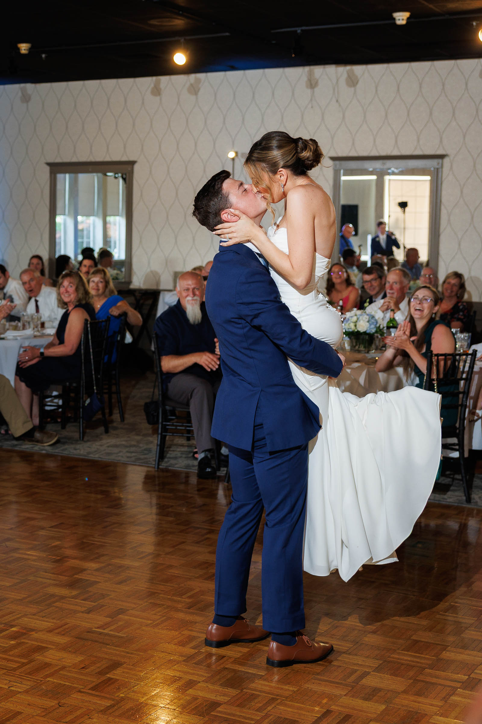 Log Cabin Wedding Photos Holyoke-21 A groom in a navy suit lifts and kisses his bride in a white dress on the dance floor, as smiling guests look on—capturing the joy of The Log Cabin Holyoke Wedding Photos.