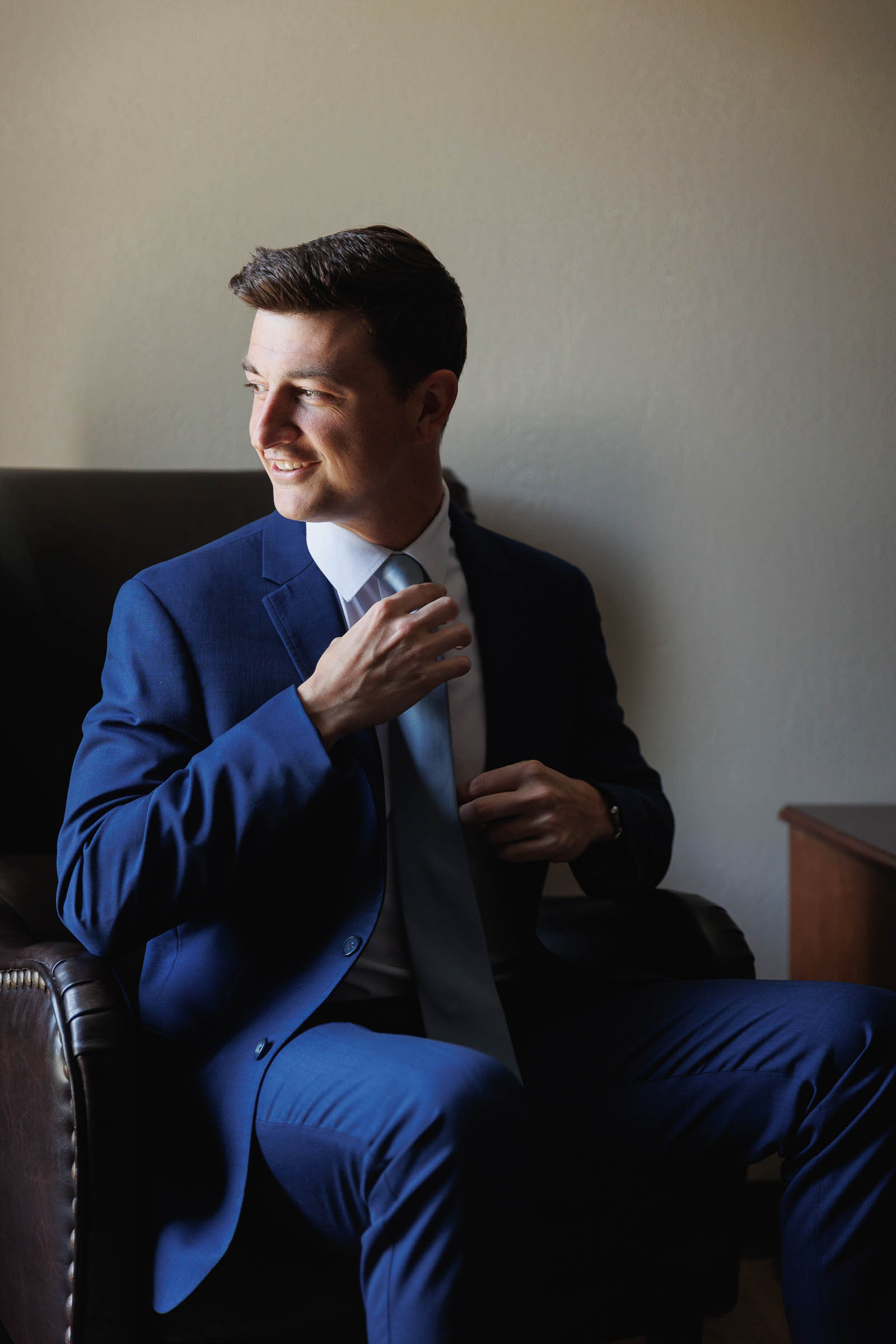 Log Cabin Wedding Photos Holyoke-6 A man in a blue suit sits on a leather chair, adjusting his tie and looking to the side as sunlight pours through the window—capturing a quiet moment reminiscent of The Log Cabin Holyoke Wedding Photos.