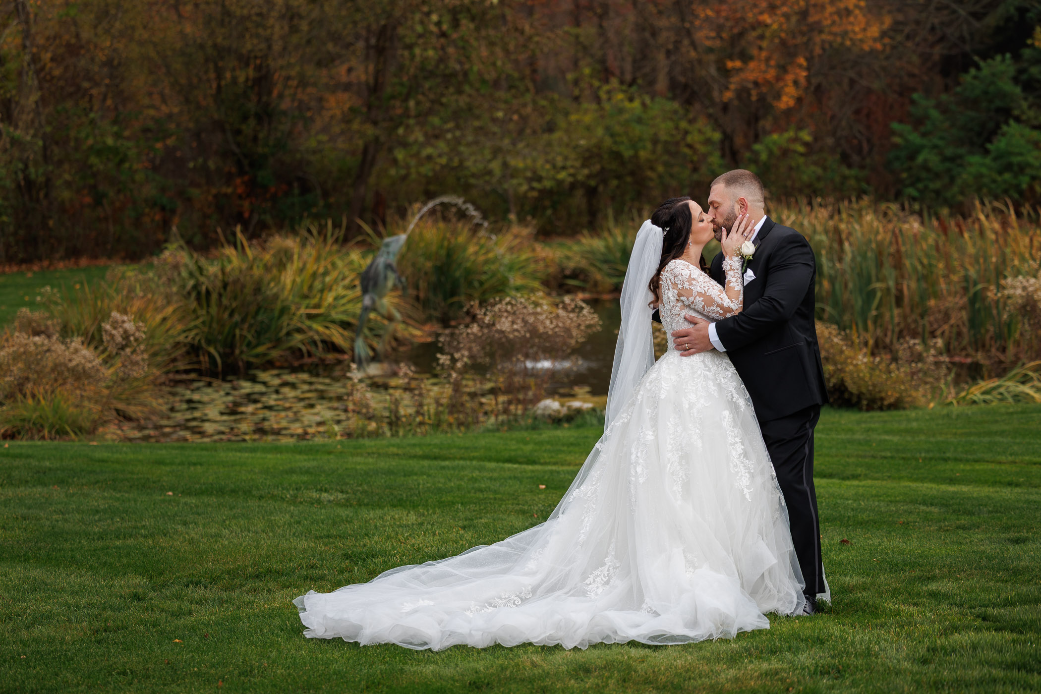 A bride and groom in formal wedding attire kiss on a grassy lawn with a pond and autumn foliage in the background, beautifully captured by The Log Cabin Wedding Photography.