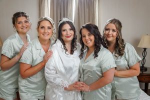 Five women pose together indoors for a Ranch Wedding Photo, with one in a white robe and tiara and the others in matching light-colored pajamas, all smiling at the camera.