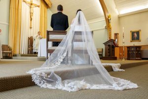 A bride and groom kneel at the altar in a church during their wedding ceremony, the bride's long veil spread across the floor in this heartfelt Ranch Wedding Photo.