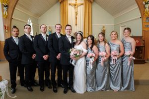 A bride and groom stand with their wedding party in formal attire inside a church, with a crucifix and altar visible in the background—a timeless moment captured as part of The Ranch Wedding Photo collection.