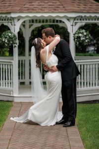 A bride and groom share a kiss in front of a white gazebo on a brick path, surrounded by greenery, creating the perfect The Ranch Wedding Photo.