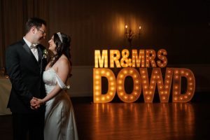 A bride and groom stand together in front of large illuminated letters spelling "MR & MRS DOWD" in a dimly lit room, capturing the magic of their special day in this unforgettable The Ranch Wedding photo.