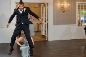 A man in a tuxedo jumps over a crouching woman in a formal dress at an indoor event, capturing a playful moment—The Ranch Wedding Photo adds an unexpected twist to the classic wedding reception.