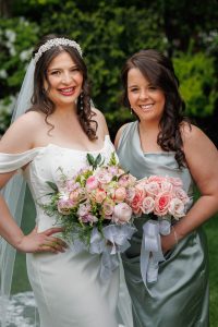 Two women stand side by side outdoors, smiling at the camera in this charming The Ranch Wedding Photo. One wears a white wedding dress and holds a bouquet; the other wears a silver dress with pink roses.