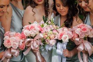 Five women in satin dresses hold bouquets of pink and white roses tied with satin ribbons, gathered closely together and looking down at the flowers—a classic moment captured by The Ranch Wedding Photo.