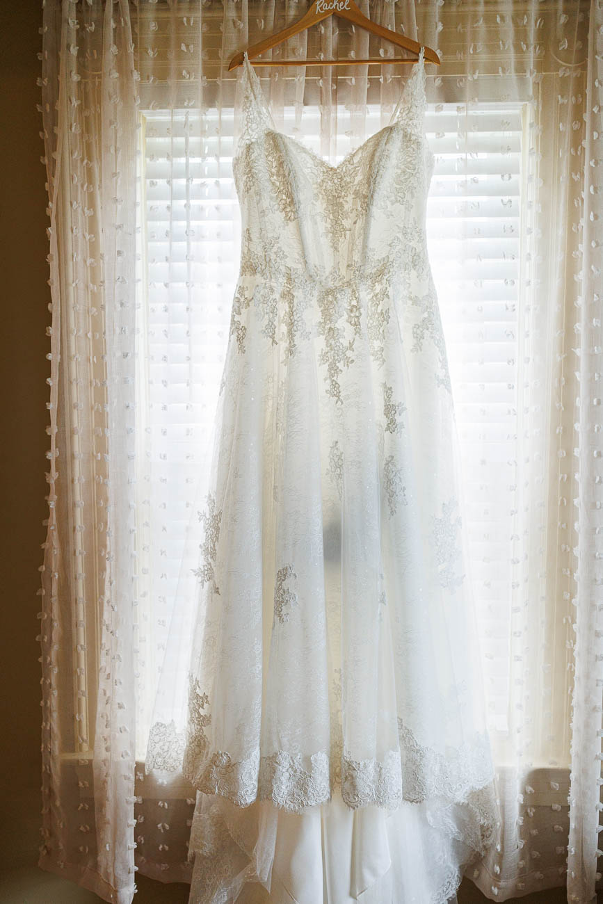The Ranch Wedding photos-1 A white lace wedding dress hangs on a wooden hanger in front of a window with sheer curtains and blinds, ready for The Ranch Wedding Photo.