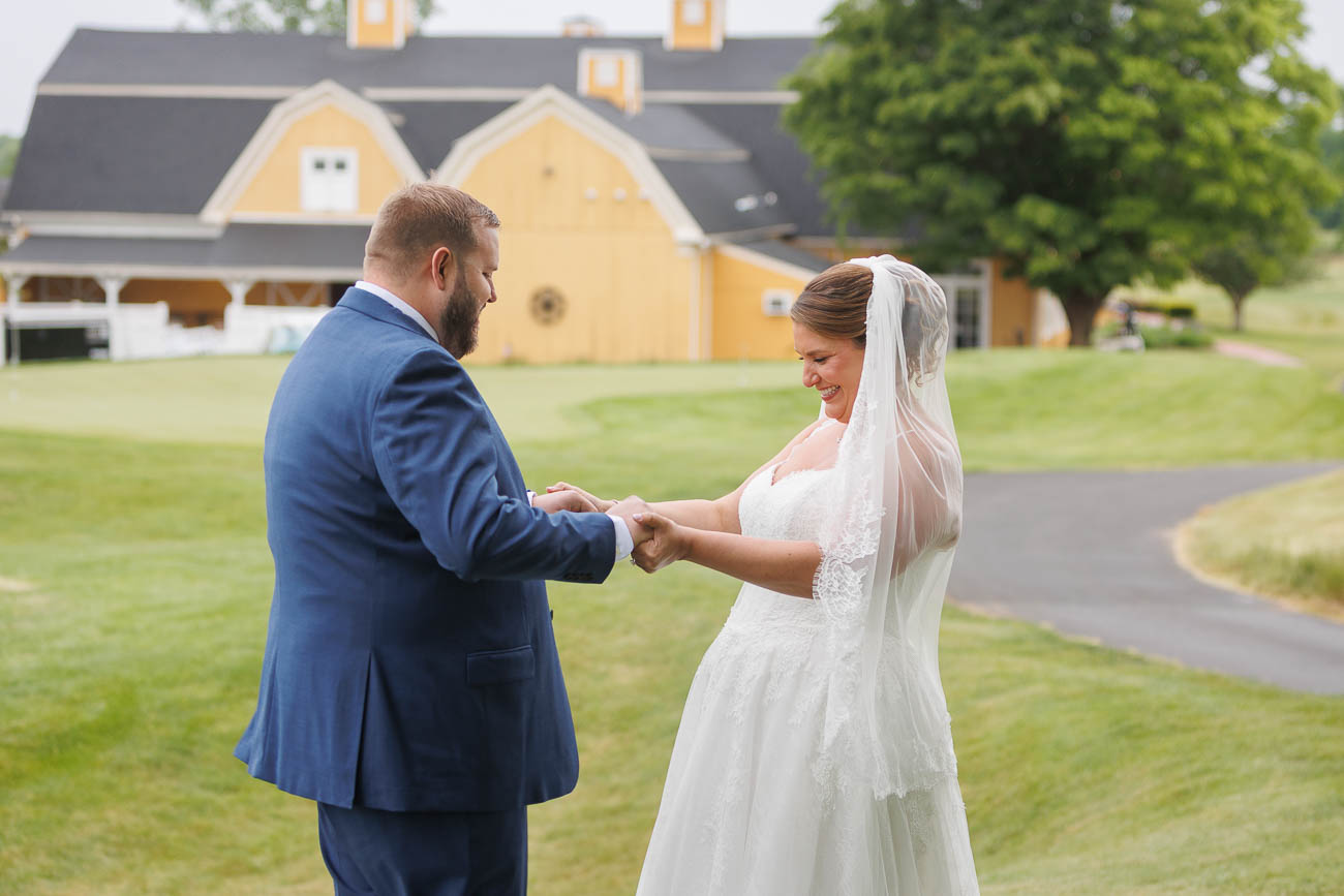 The Ranch Wedding photos-12 A bride and groom hold hands and smile at each other outdoors in front of a yellow barn on a grassy lawn, capturing the perfect Ranch Wedding Photo.