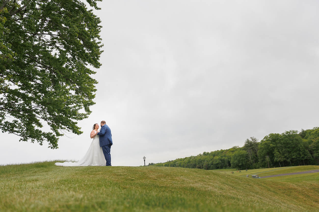 The Ranch Wedding photos-13 The Ranch Wedding Photo captures a bride and groom standing close together on a grassy hill under an overcast sky, with trees in the background.