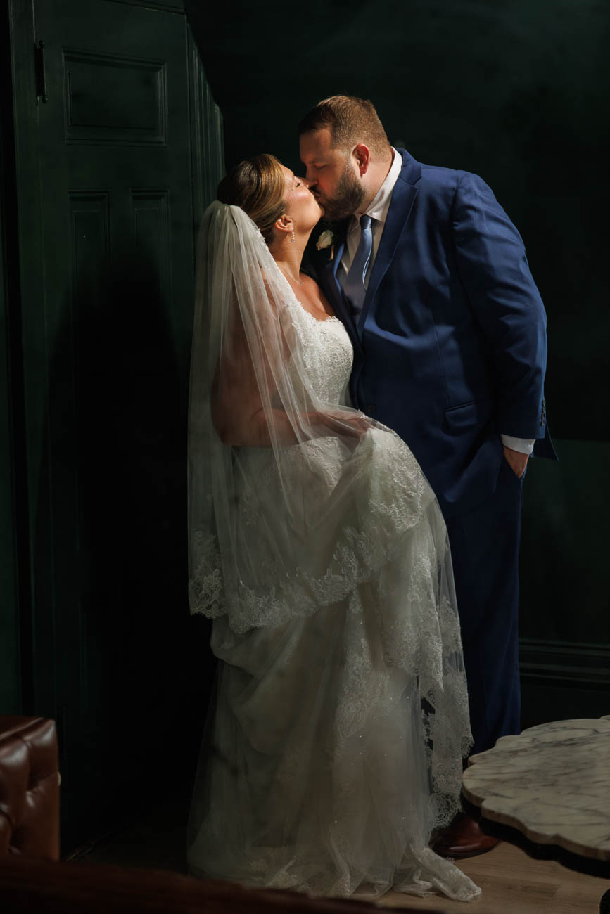 The Ranch Wedding photos-14 A bride and groom in formal wedding attire share a kiss indoors at The Ranch Wedding Photo, with the bride in a white dress and veil, and the groom in a blue suit.