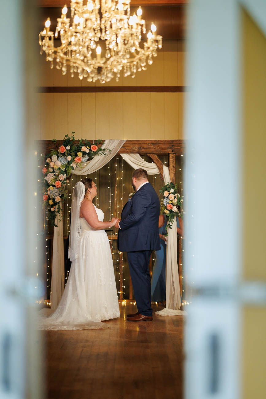 The Ranch Wedding photos-15 A bride and groom stand under a floral arch during their wedding ceremony, holding hands and facing each other beneath a chandelier—an unforgettable moment captured in The Ranch Wedding Photo.