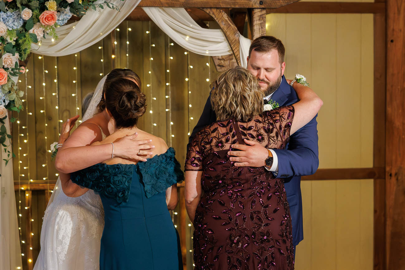 The Ranch Wedding photos-16 Two couples embrace during a wedding ceremony at The Ranch Wedding Photo, with floral decorations and string lights in the background.