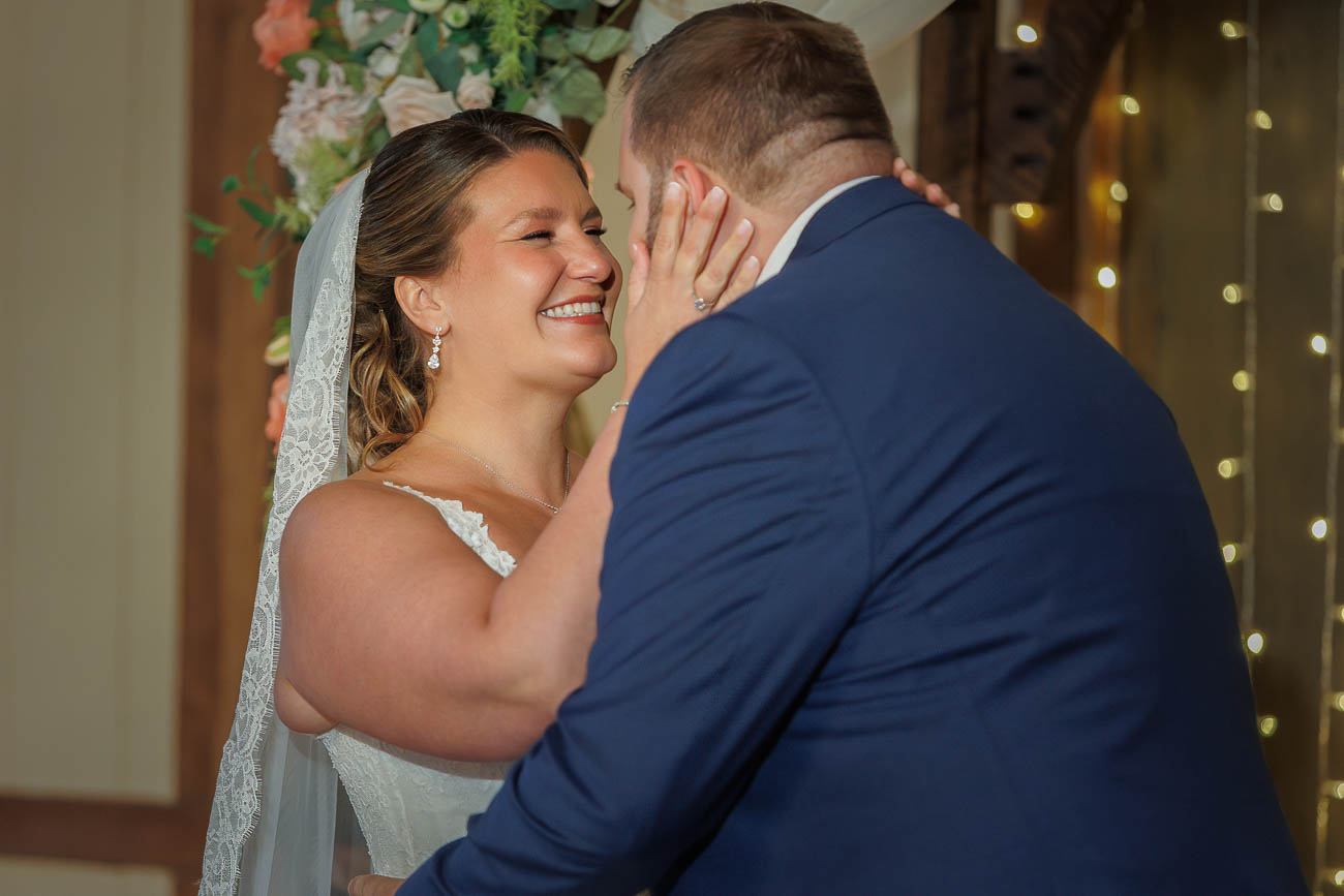 The Ranch Wedding photos-17 A bride and groom face each other and smile during their wedding ceremony at The Ranch, with the bride gently touching the groom's face. Floral decorations and string lights complete this unforgettable Ranch Wedding Photo setting.