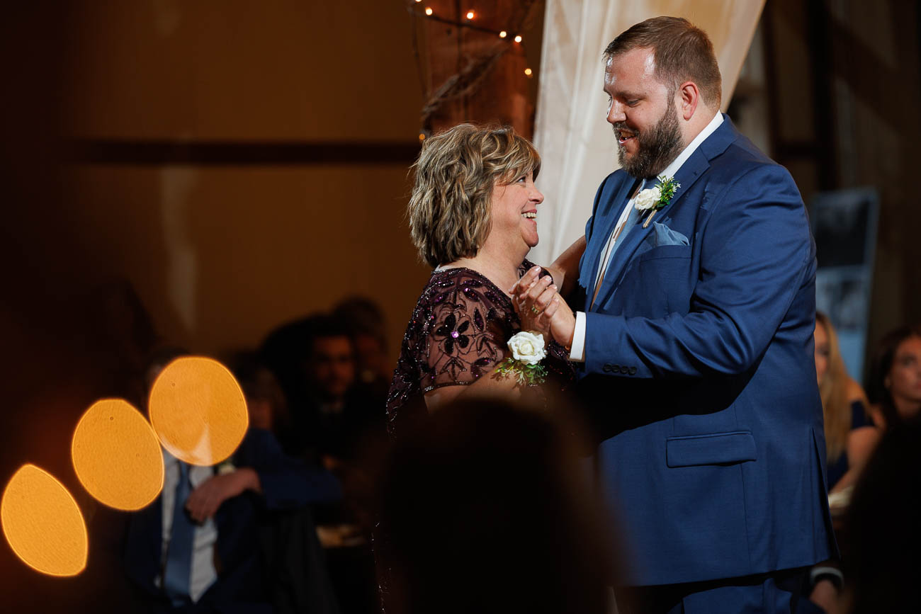 The Ranch Wedding photos-19 A man in a blue suit and a woman in a dark dress with a corsage share a dance at The Ranch Wedding Photo, smiling at each other under warm lighting at this formal indoor event.