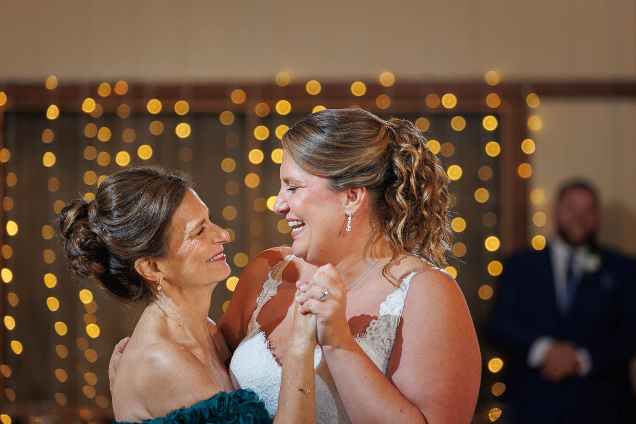 The Ranch Wedding photos-20 Two women in formal attire, one in a wedding dress, smile and hold hands while dancing beneath string lights at The Ranch Wedding Photo backdrop.