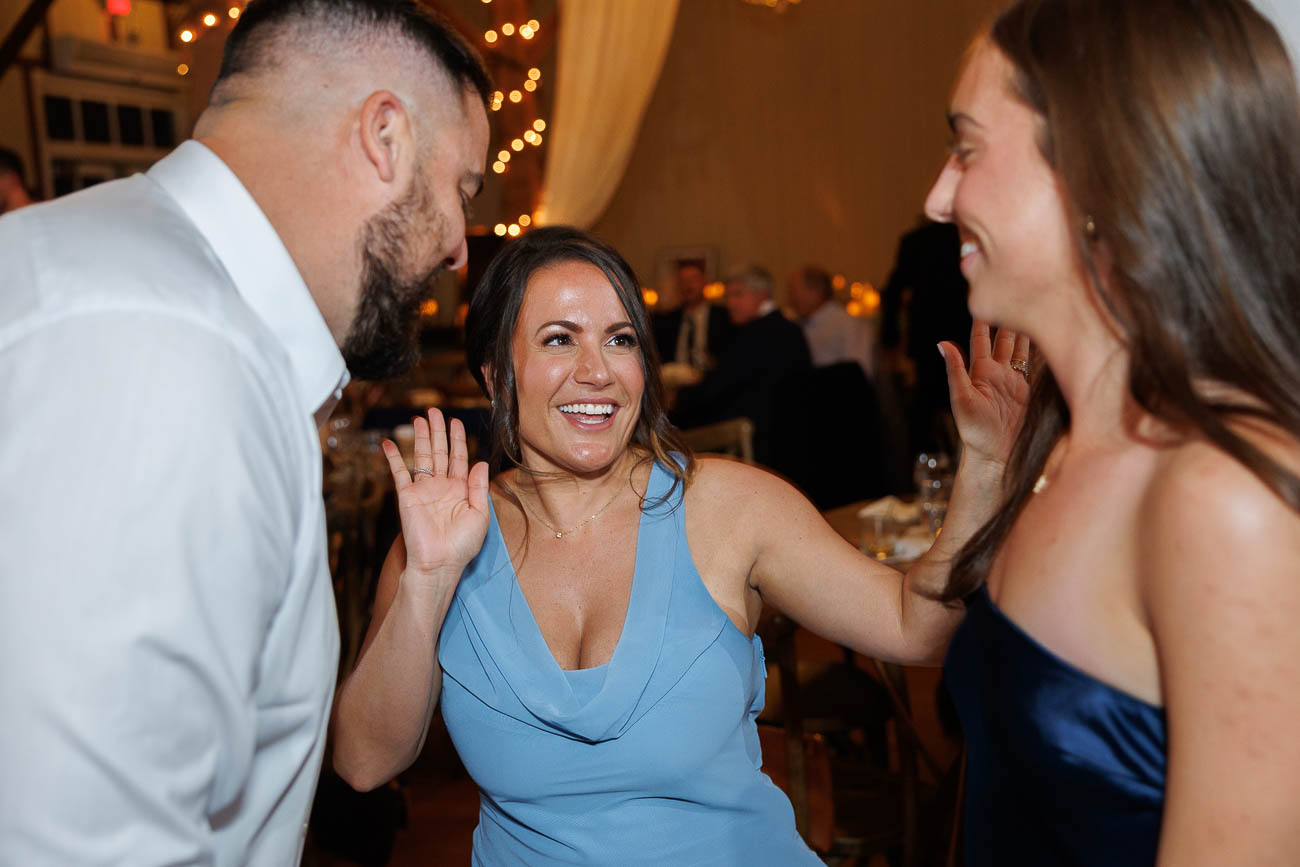 The Ranch Wedding photos-21 Three adults dressed in formal attire smile and interact with each other at an indoor event, illuminated by warm lighting and string lights in the background—captured perfectly as a The Ranch Wedding Photo.