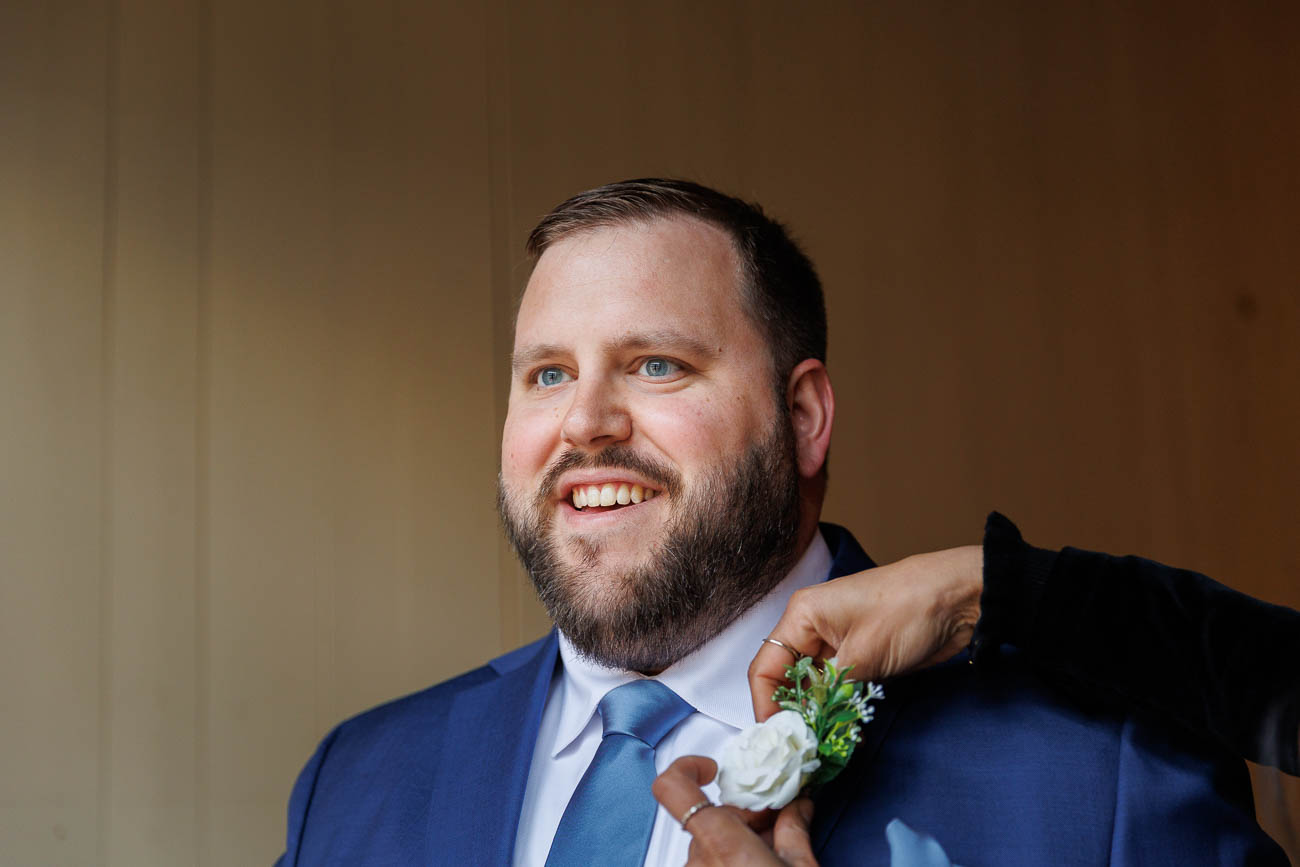 The Ranch Wedding photos-3 A man in a blue suit smiles as someone adjusts a boutonniere on his lapel in this charming The Ranch Wedding Photo.