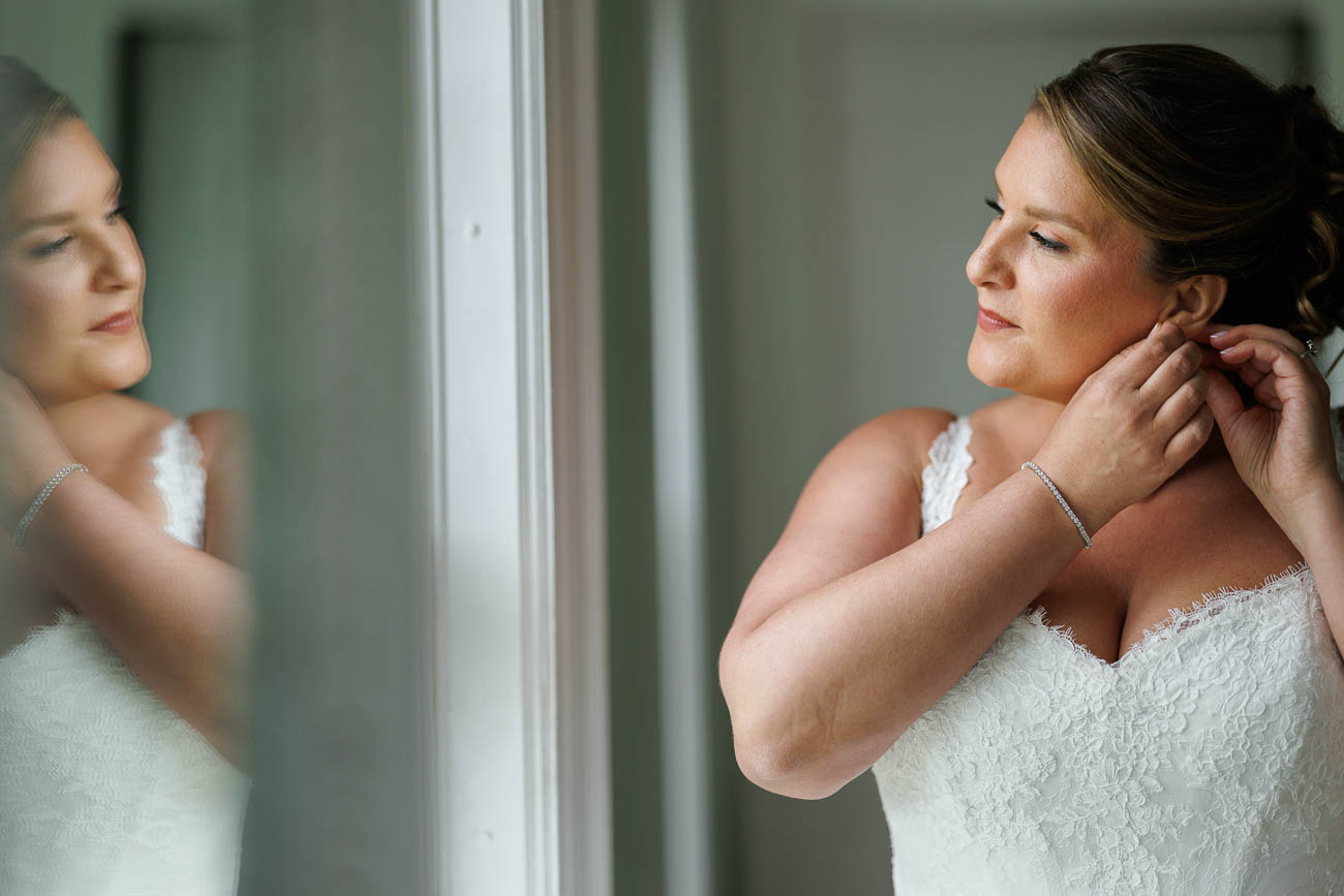 The Ranch Wedding photos-9 A woman in a white lace wedding dress stands by a window at The Ranch, adjusting her earring and gazing at her reflection in the glass—a stunning Ranch Wedding Photo moment.