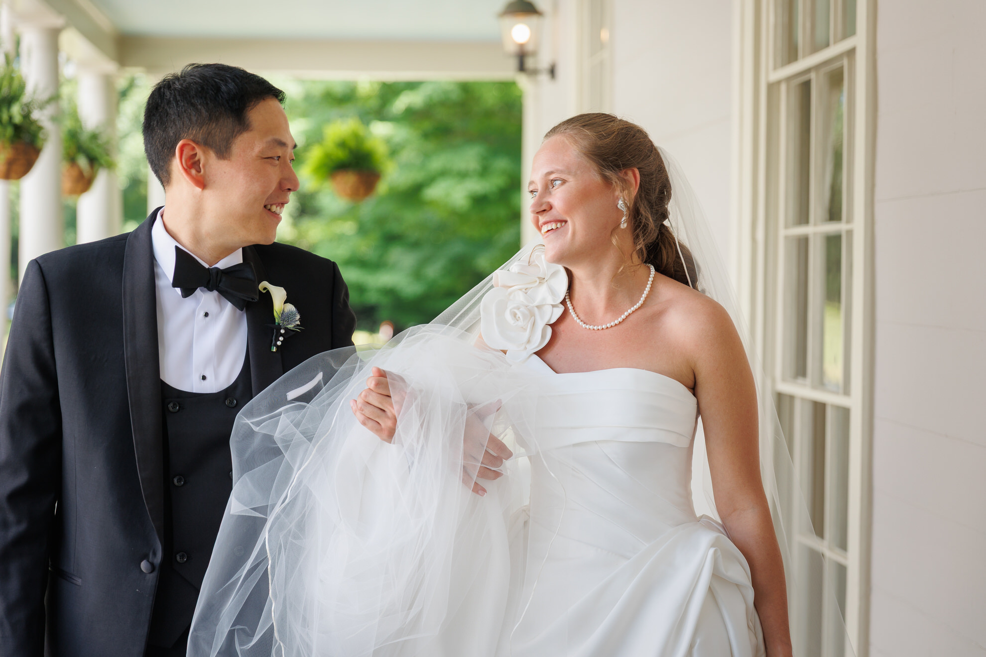 Natasha and Fang-119 Lyman Estate Wedding A bride in a white dress and a groom in a tuxedo smile at each other while standing on a porch with hanging plants during their Lyman Estates Wedding.