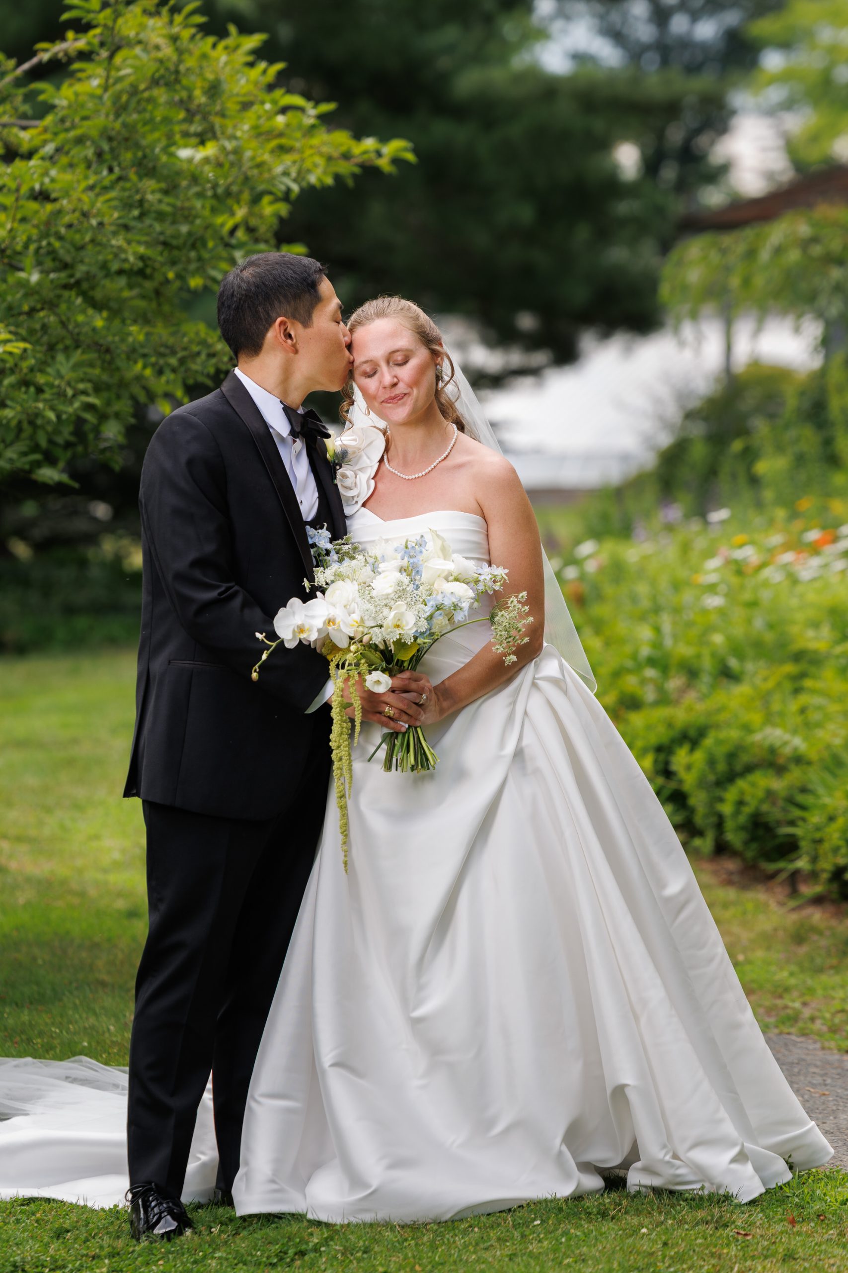 Natasha and Fang-177 Lyman Estate Wedding A groom in a black suit kisses a bride in a white dress holding a bouquet, standing together outdoors in the beautiful garden setting of their Lyman Estates Wedding.