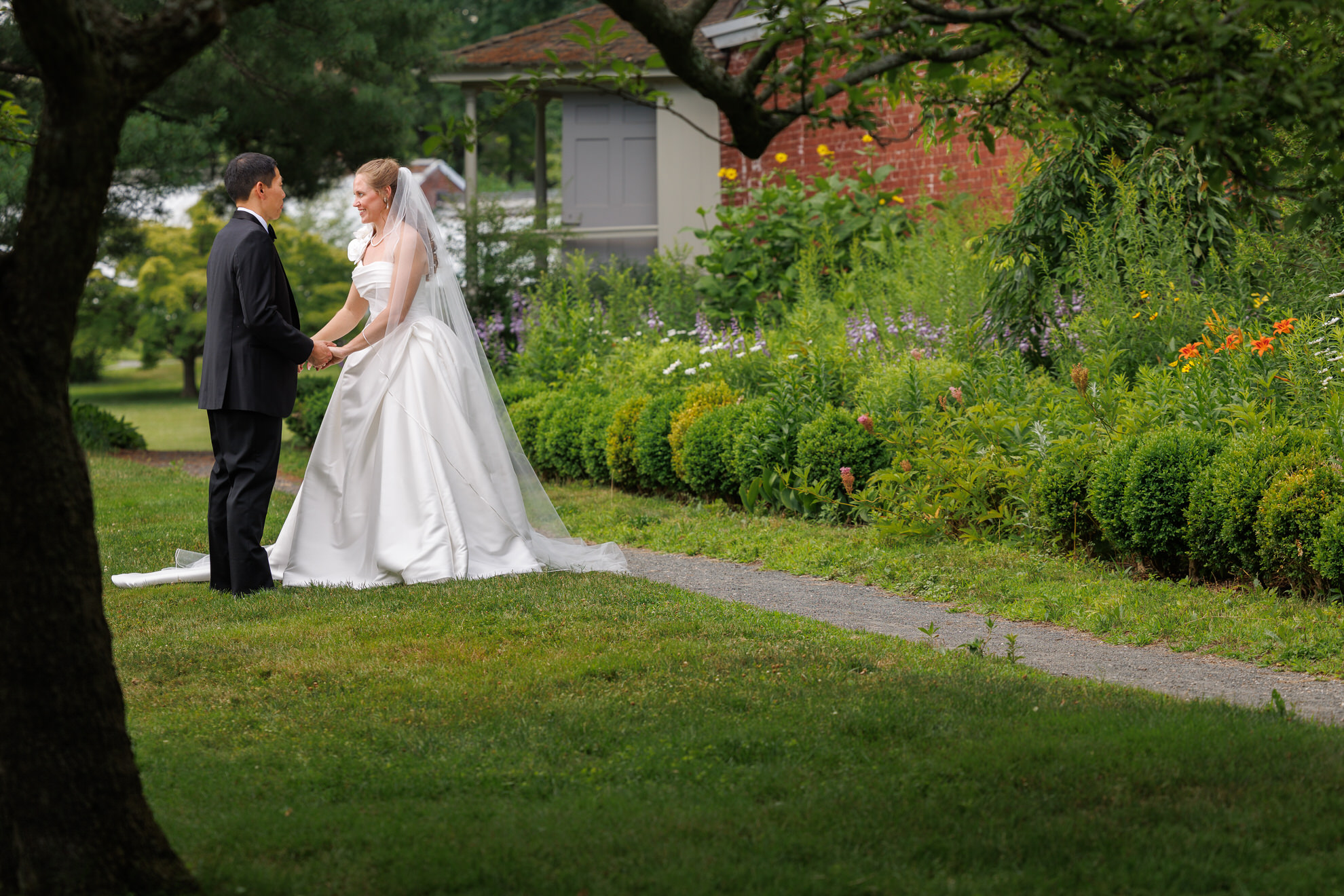 Natasha and Fang-189 Lyman Estate Wedding A bride and groom stand holding hands on a grassy lawn near a garden, with trees and the historic Lyman Estate building in the background, capturing the timeless elegance of a Lyman Estates Wedding.