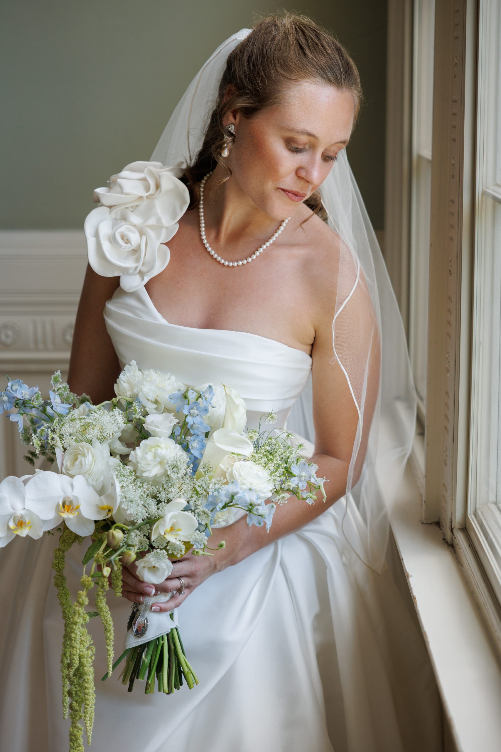 Natasha and Fang-209 Lyman Estate Wedding Bride in a white dress and veil holds a bouquet of white and light blue flowers, standing by a window and looking down during her Lyman Estates Wedding.