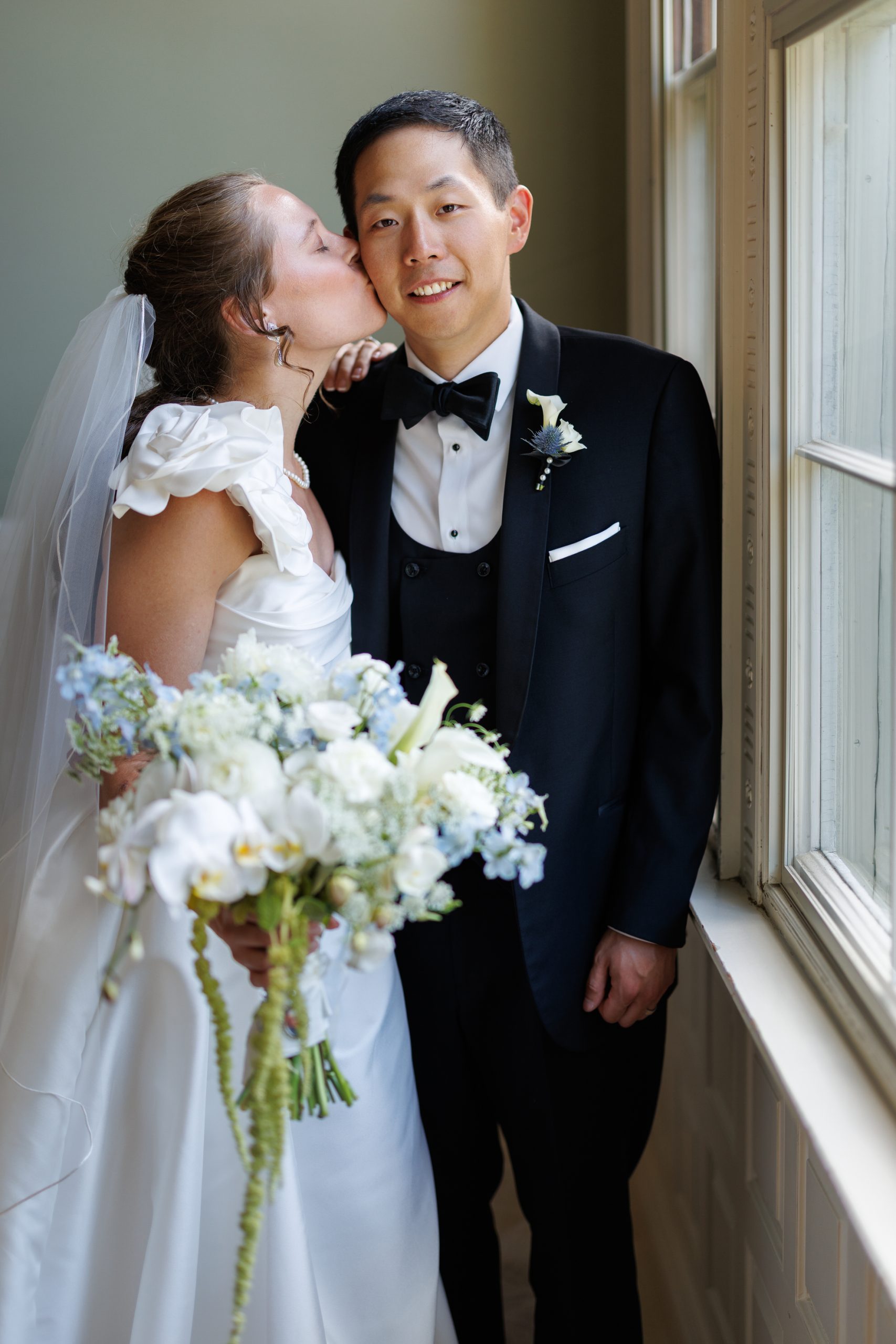 Natasha and Fang-220 Lyman Estate Wedding Bride in a white dress kisses groom in a black tuxedo on the cheek while holding a bouquet, standing by a window at their romantic Lyman Estates wedding.