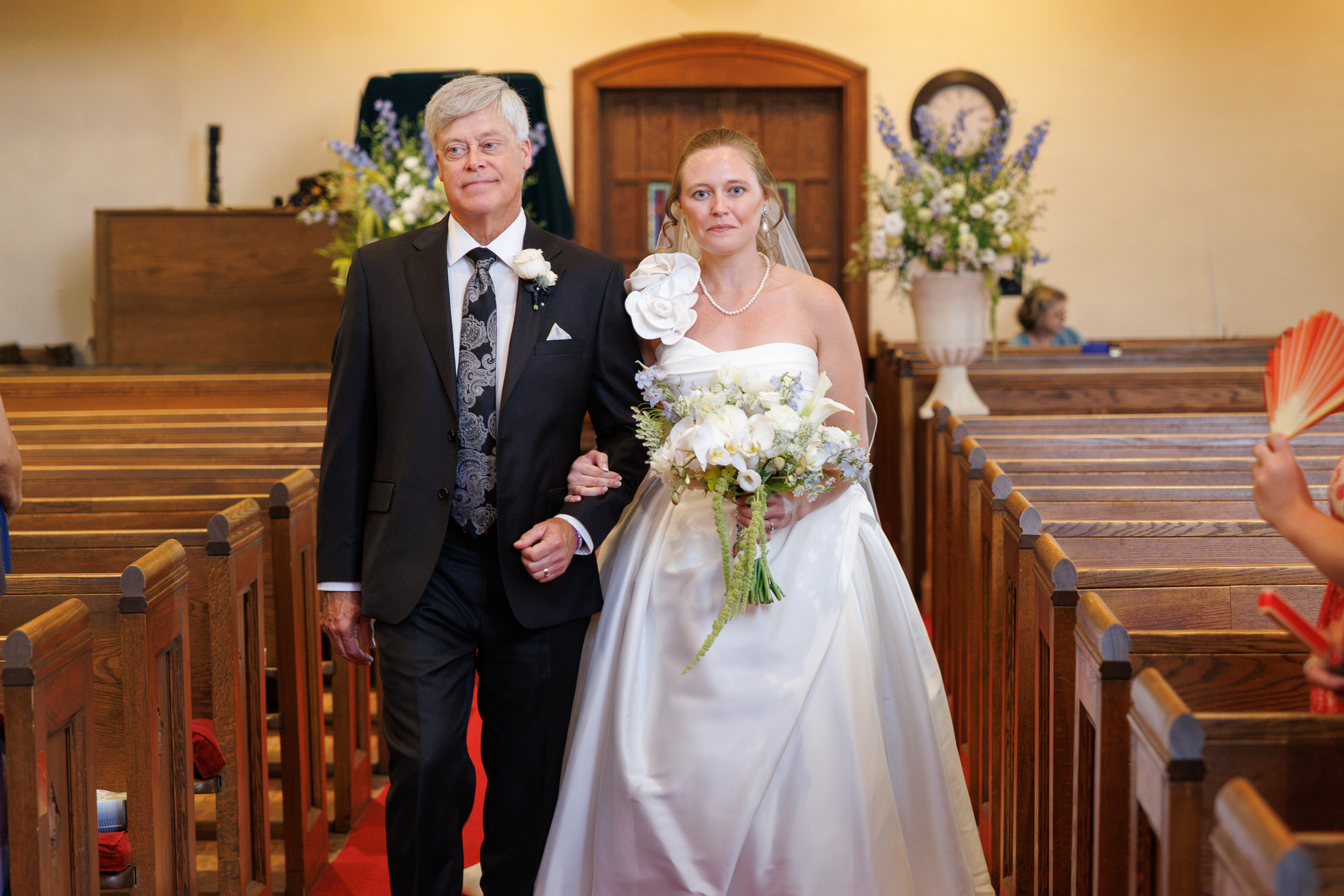 Natasha and Fang-285 Lyman Estate Wedding A bride in a white dress holding flowers walks down the aisle with an older man in a suit during a charming Lyman Estates Wedding, surrounded by wooden pews and beautiful flower arrangements inside the church.