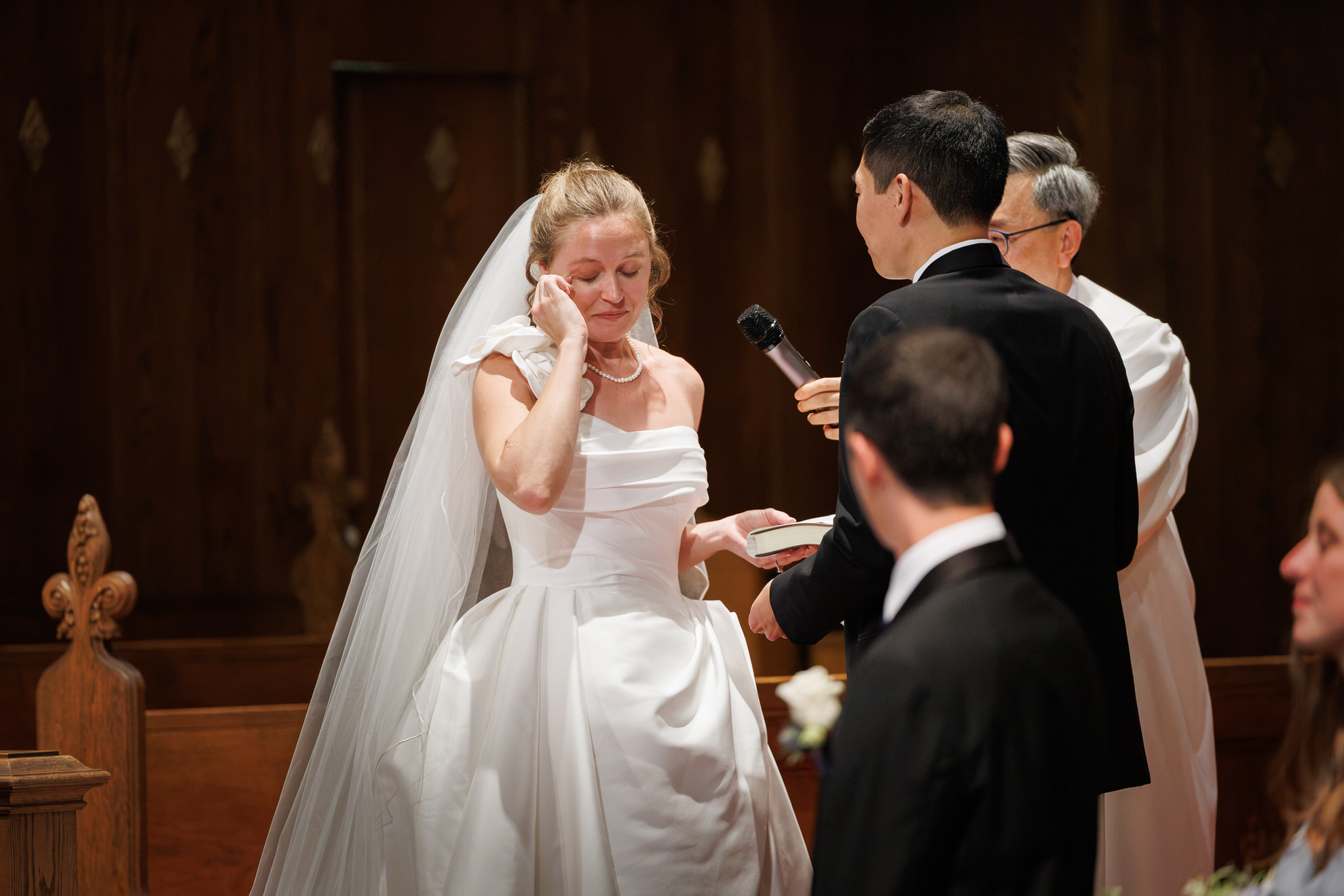 Natasha and Fang-347 Lyman Estate Wedding A bride in a white dress wipes a tear while standing beside the groom during their Lyman Estates Wedding ceremony, with an officiant and guests nearby.