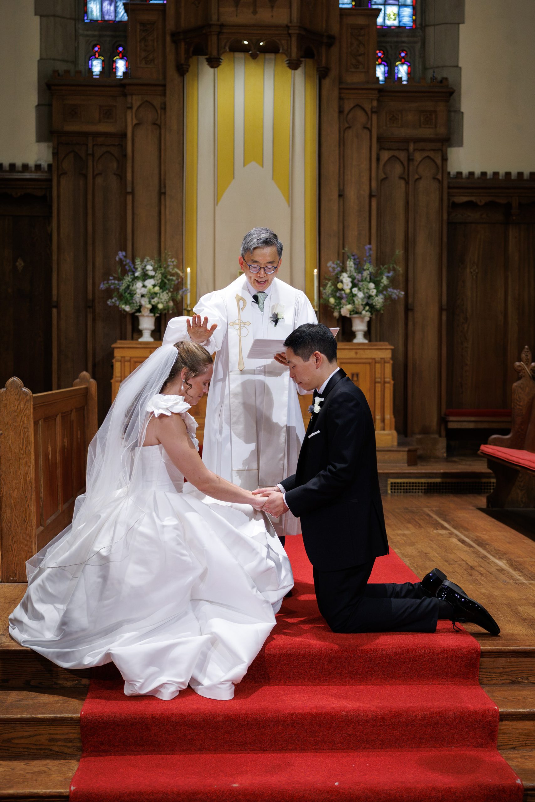 Natasha and Fang-381 Lyman Estate Wedding A bride and groom kneel together on a red carpet in a church while an officiant in white robes performs a wedding ceremony, capturing the timeless elegance reminiscent of a Lyman Estates Wedding.