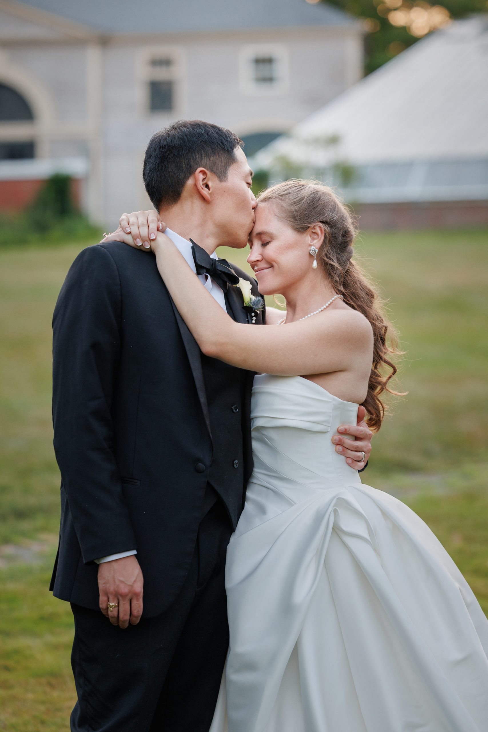 Natasha and Fang-600 Lyman Estate Wedding A groom in a black suit kisses a bride in a white dress on the forehead as they embrace outdoors in front of a building during their elegant Lyman Estates Wedding.