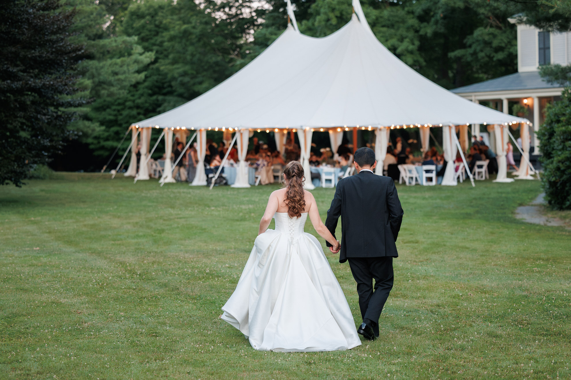 Natasha and Fang-626 Lyman Estate Wedding A bride and groom walk hand in hand across grass toward a white tent filled with seated guests at their elegant Lyman Estates wedding reception.