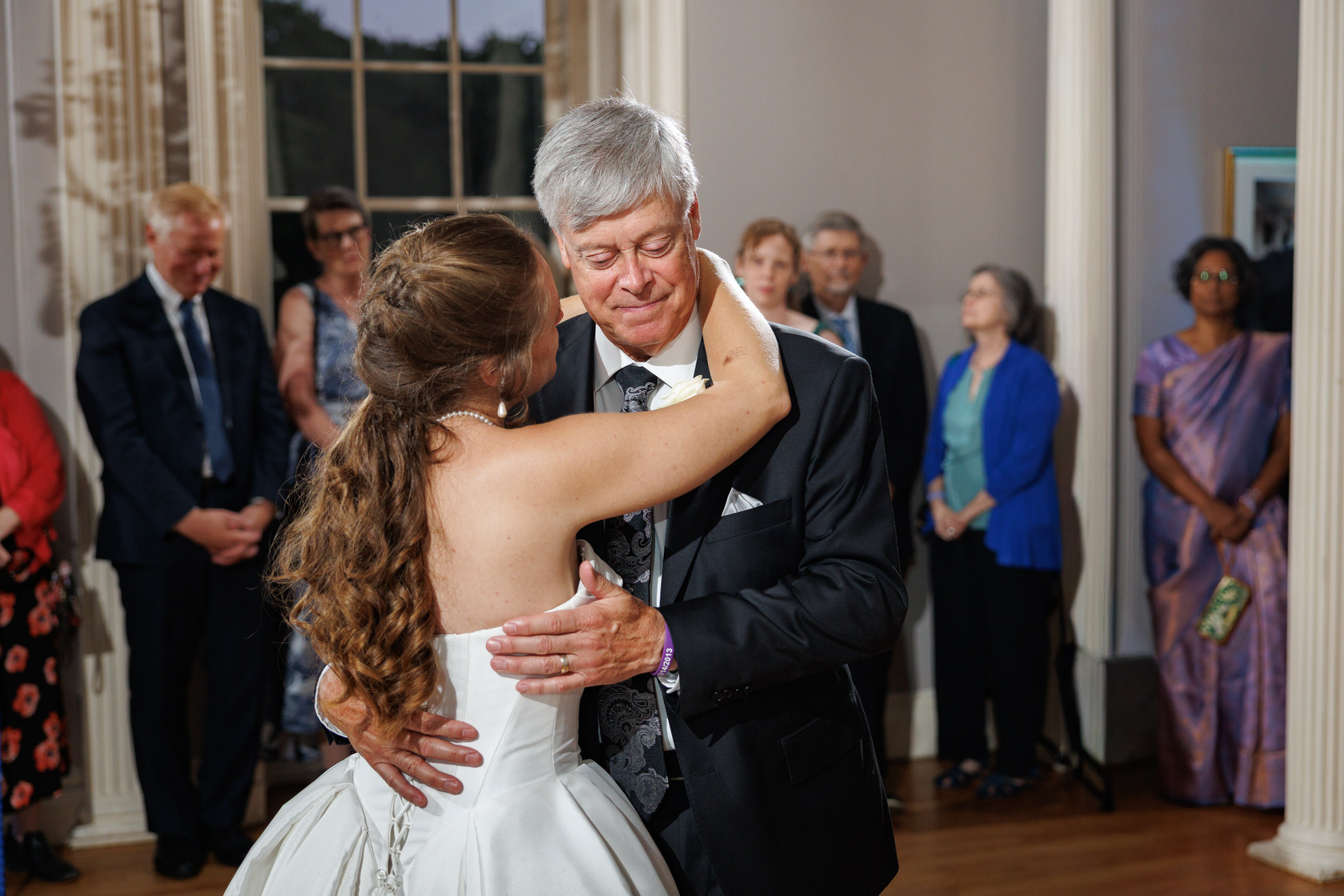 Natasha and Fang-676 Lyman Estate Wedding A bride in a white dress dances with an older man in a suit, surrounded by formally dressed guests in a well-lit room at their elegant Lyman Estates wedding.