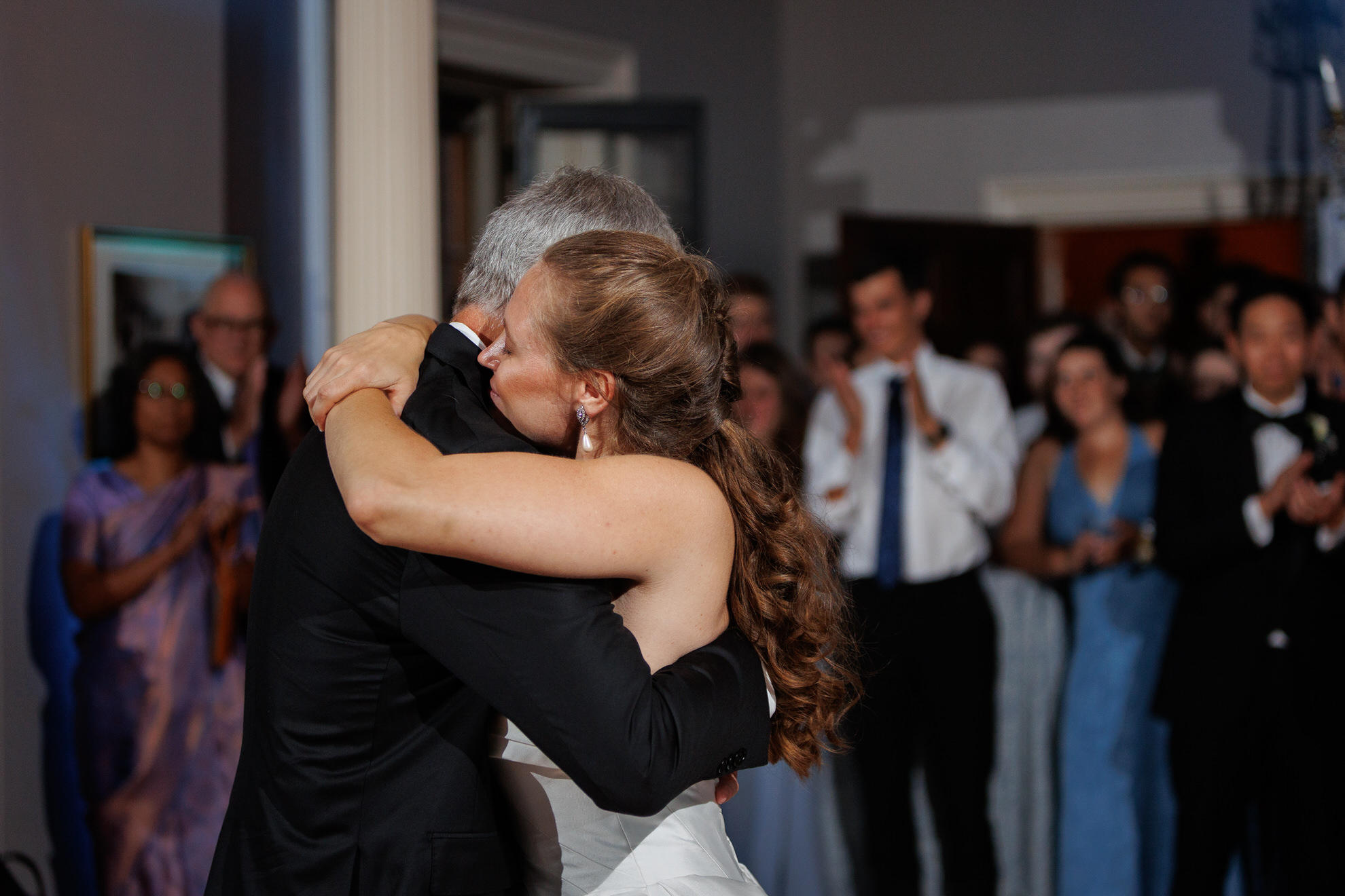 Natasha and Fang-685 Lyman Estate Wedding A bride in a white dress hugs an older man in a suit on the dance floor during a Lyman Estates Wedding, while formally dressed guests watch and applaud in the background.