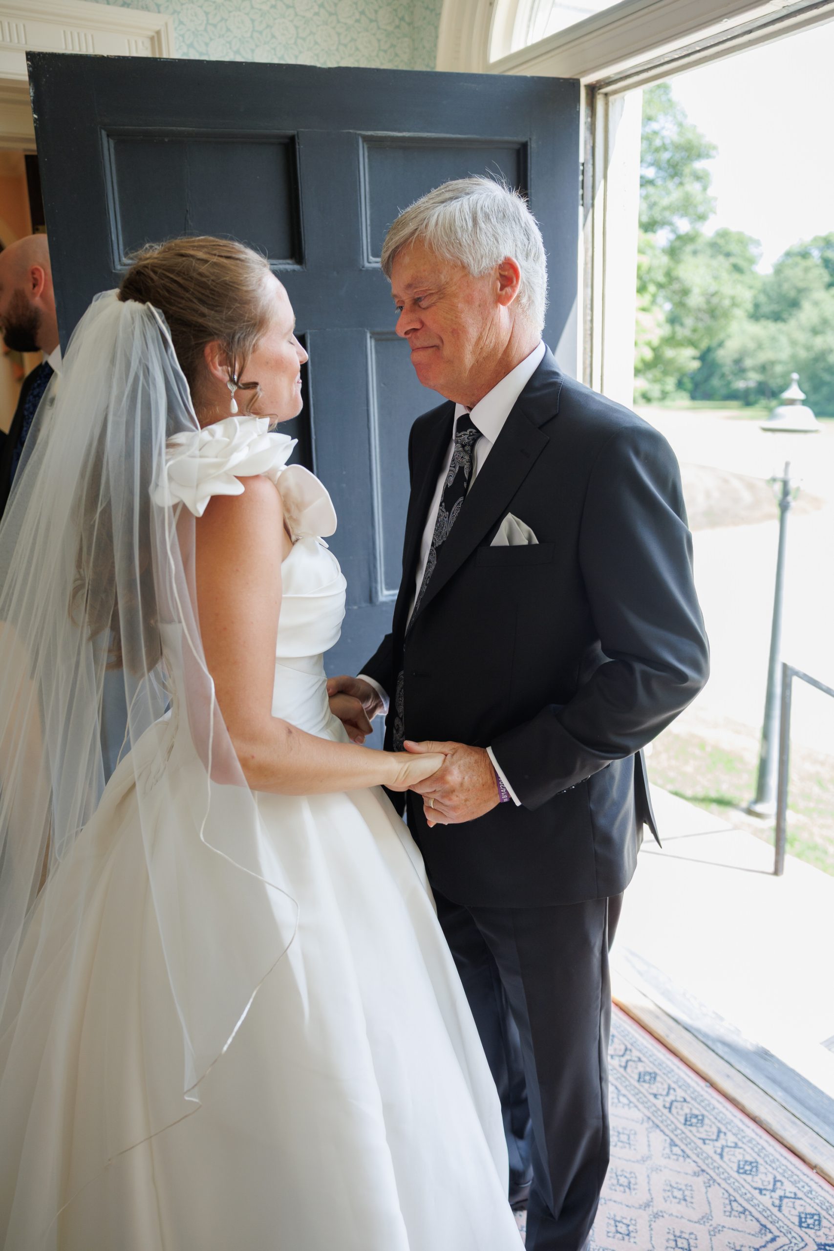 Natasha and Fang-74 Lyman Estate Wedding A bride in a white gown and veil holds hands and smiles with an older man in a suit, standing together by an open door with sunlight outside at their Lyman Estates Wedding.