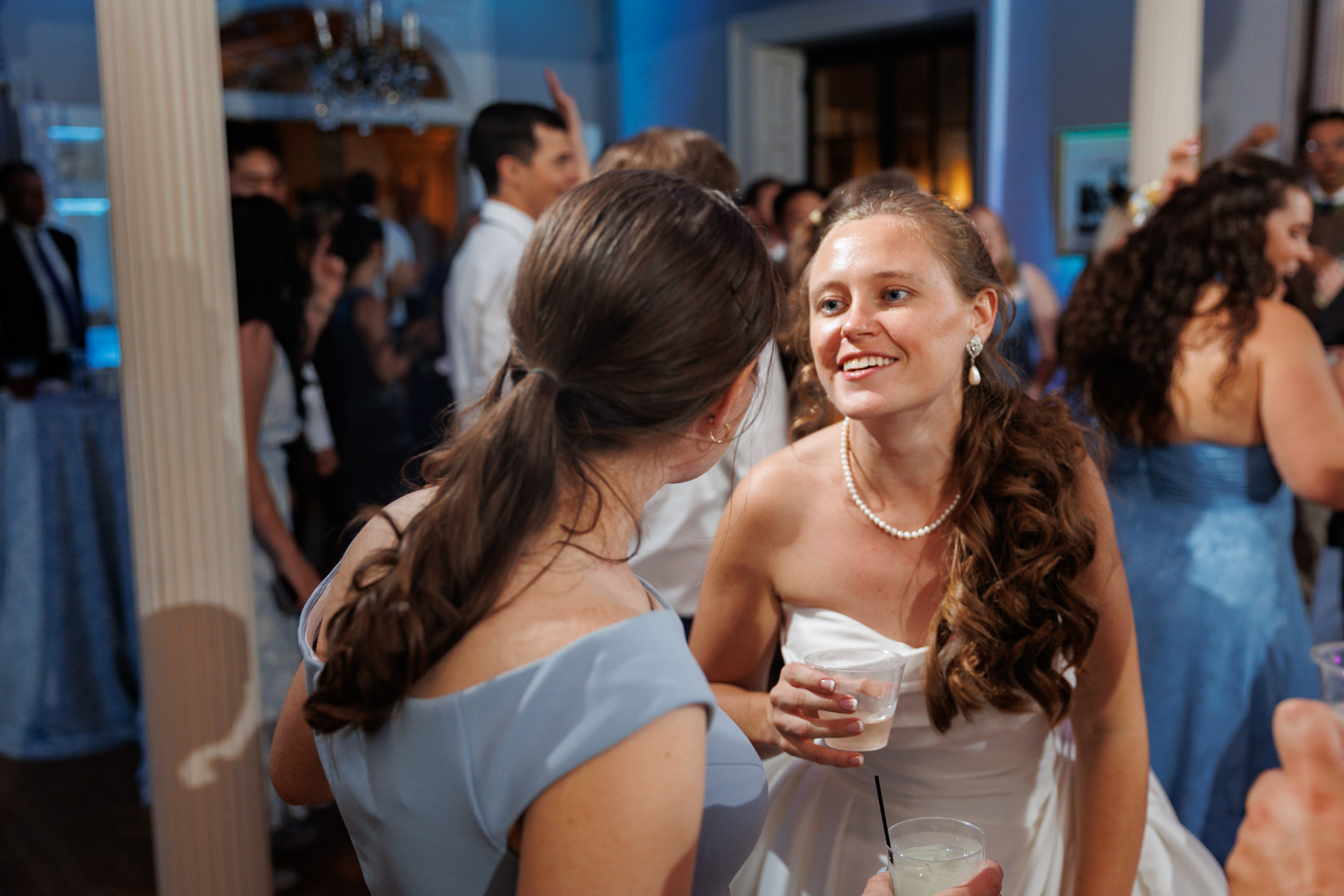 Natasha and Fang-741 Lyman Estate Wedding At a lively Lyman Estates Wedding, a woman in a white dress holding a drink talks to another woman in a blue dress at the crowded indoor event.