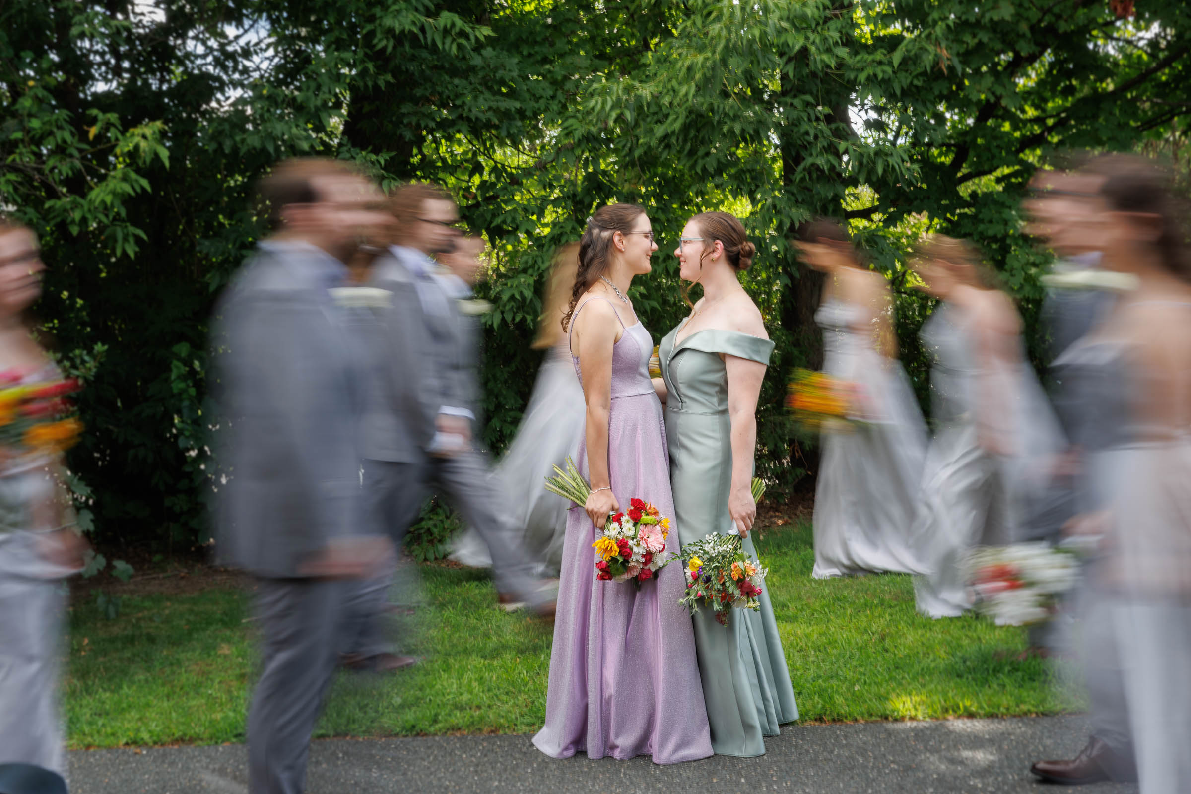 Two women in formal dresses face each other holding bouquets, while wedding party members in motion blur walk past them outdoors.