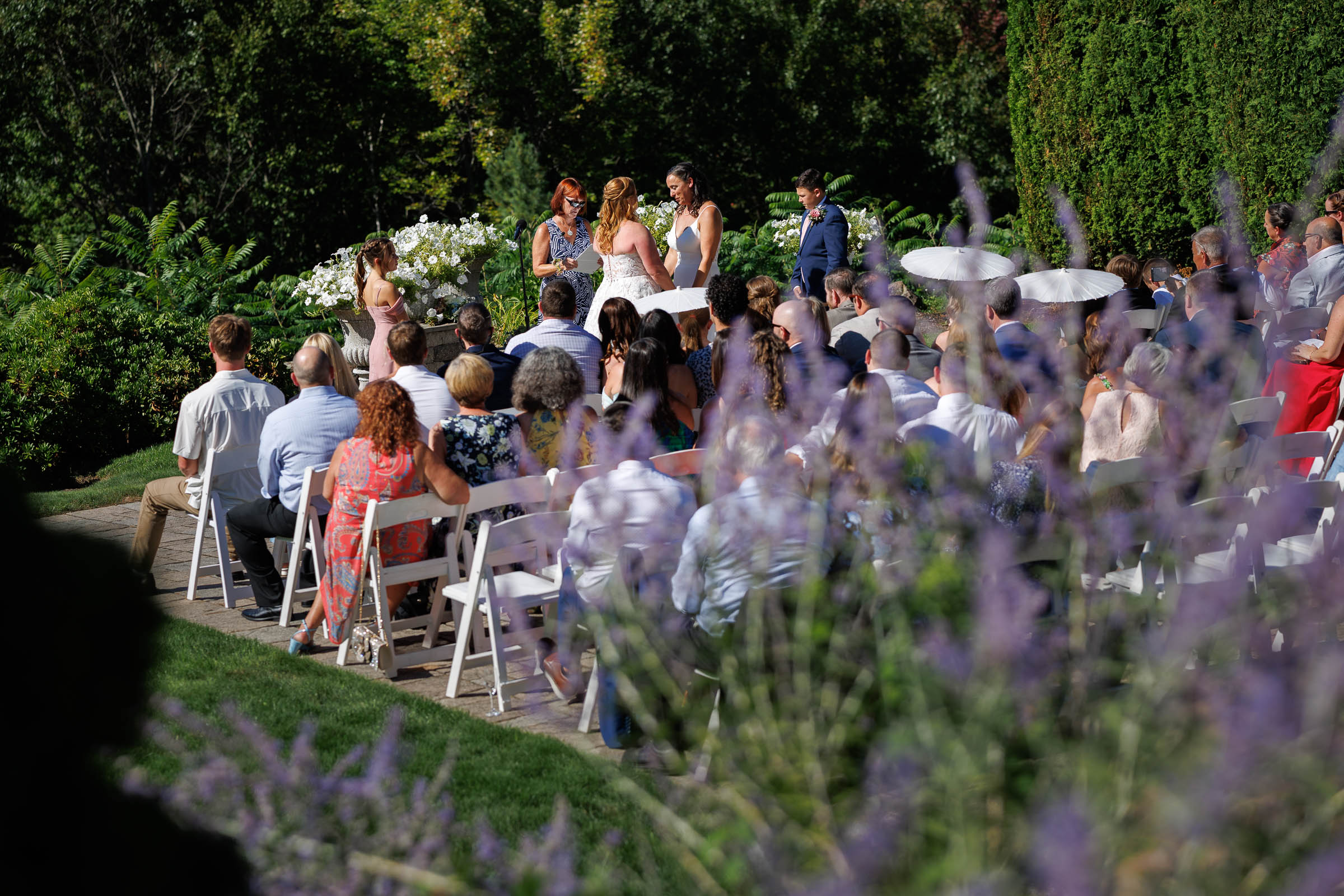 Guests seated outdoors at the Log Cabin Holyoke watch a beautiful wedding ceremony with the wedding party standing in front; purple flowers blur the foreground.