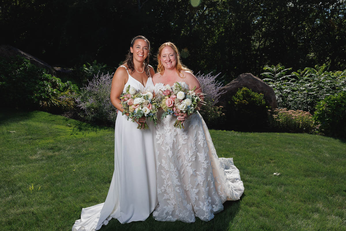 Two women in wedding dresses stand outdoors on grass, smiling at the camera and holding bouquets of flowers, with greenery and sunlight in the background—a joyful moment beautifully captured by The Log Cabin Holyoke photo and video.