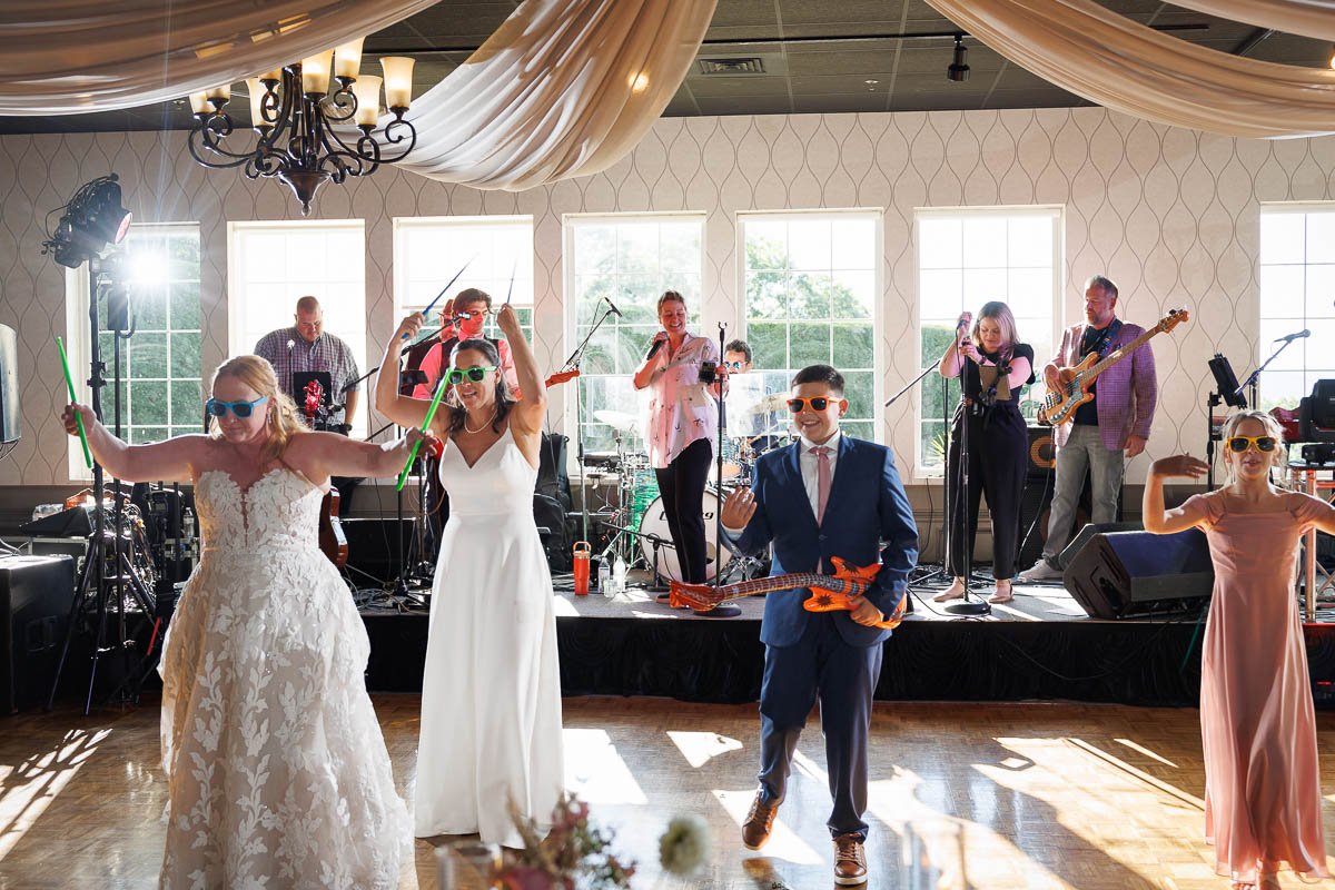 A bride, groom, and wedding party wearing sunglasses dance with toy guitars in front of a live band during a daytime wedding reception at The Log Cabin Holyoke, captured perfectly by photo and video.