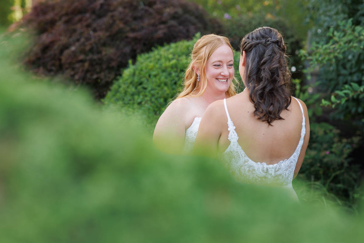 Two women in white dresses stand facing each other outdoors at The Log Cabin Holyoke, smiling, surrounded by green foliage—captured beautifully in photo and video.