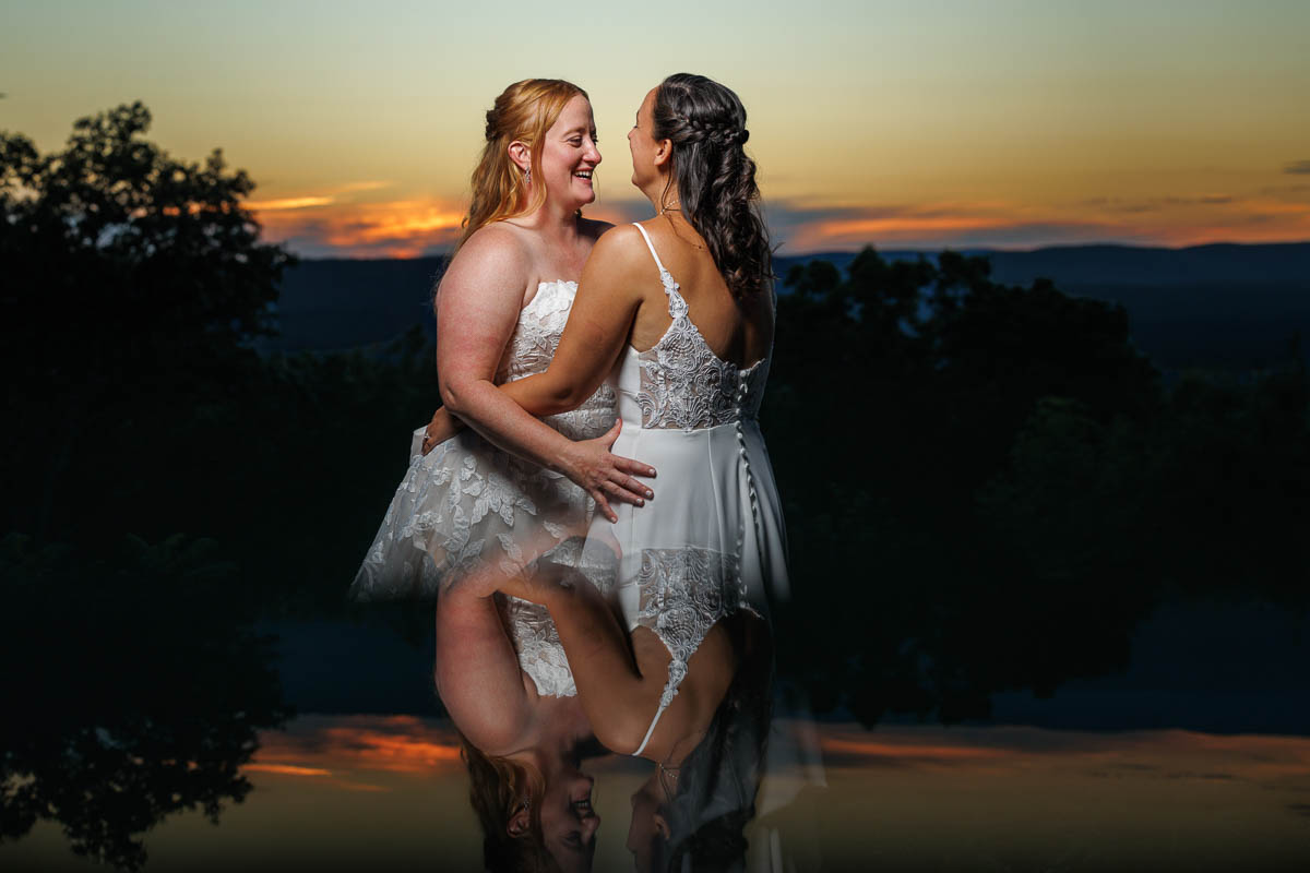 Two women in white dresses embrace and smile at each other outdoors at sunset, their reflections visible below them in this beautiful The Log Cabin Holyoke photo and video moment.