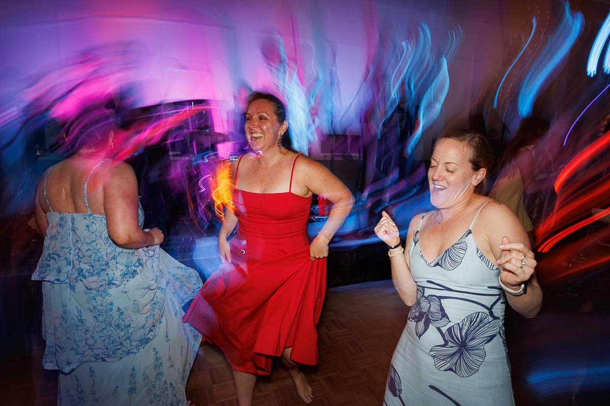 Three women dance joyfully on a wooden floor at The Log Cabin Holyoke, surrounded by colorful, blurred lights, creating a lively and energetic atmosphere captured beautifully in photo and video.