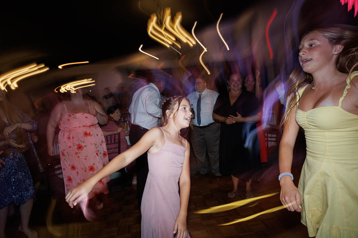 Two girls in dresses dance at a party with blurred lights and people in the background, creating a lively and energetic atmosphere captured perfectly by The Log Cabin Holyoke photo and video.