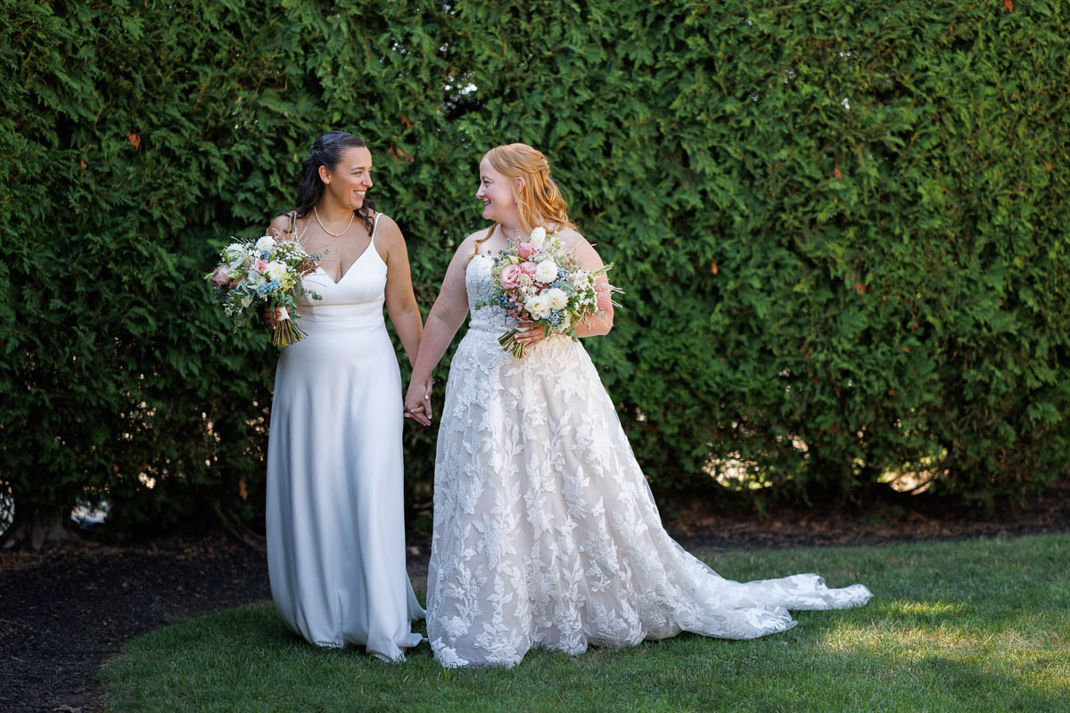 Two women in wedding dresses stand on grass holding hands and bouquets, smiling at each other, with green bushes in the background—a beautiful moment captured by The Log Cabin Holyoke photo and video.