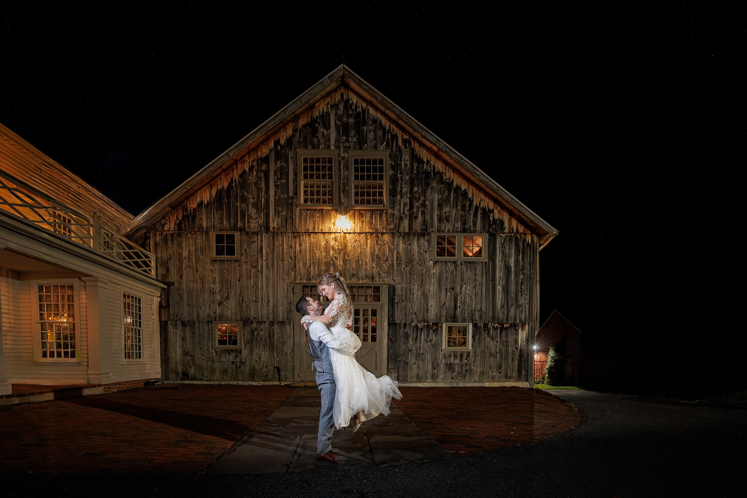 A groom lifts his bride in front of a rustic wooden barn at night, captured by a Central Mass wedding photographer.